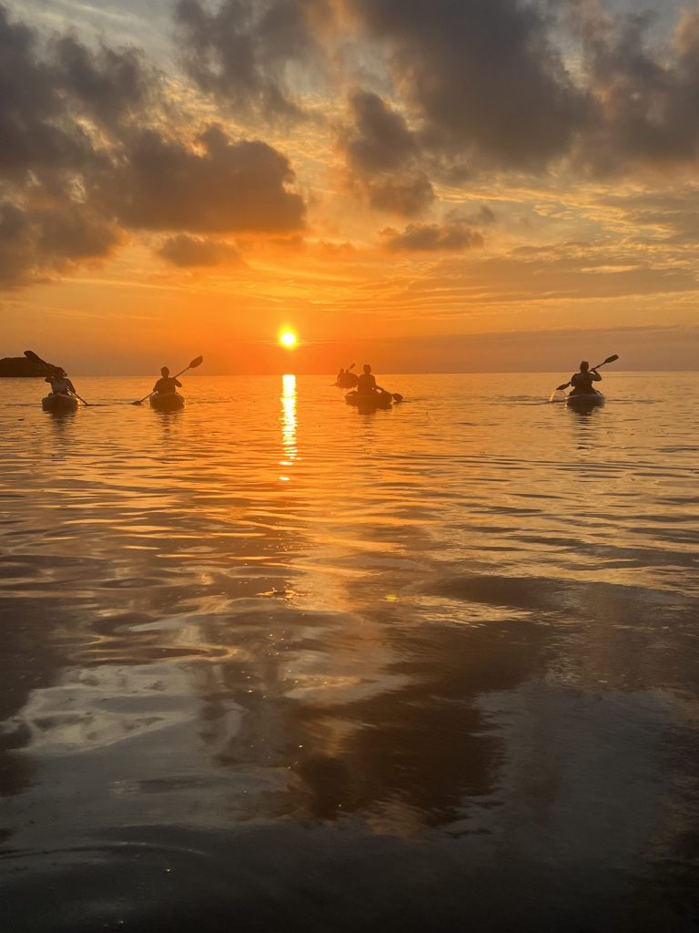 Kayakers on calm water at sunset with silhouetted clouds.