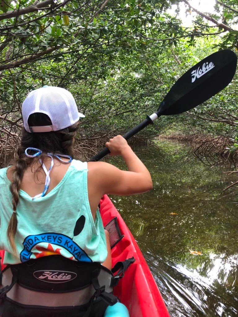 Woman kayaking through mangrove tunnel, wearing a cap and tank top.