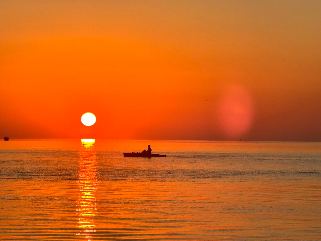 Person kayaking on calm water at sunset with the sun reflecting on the sea.