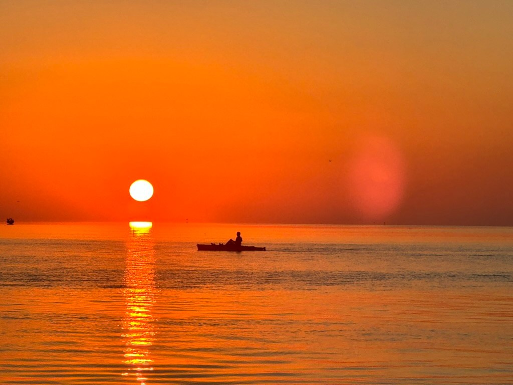 Person in kayak on calm sea during vibrant orange sunset.