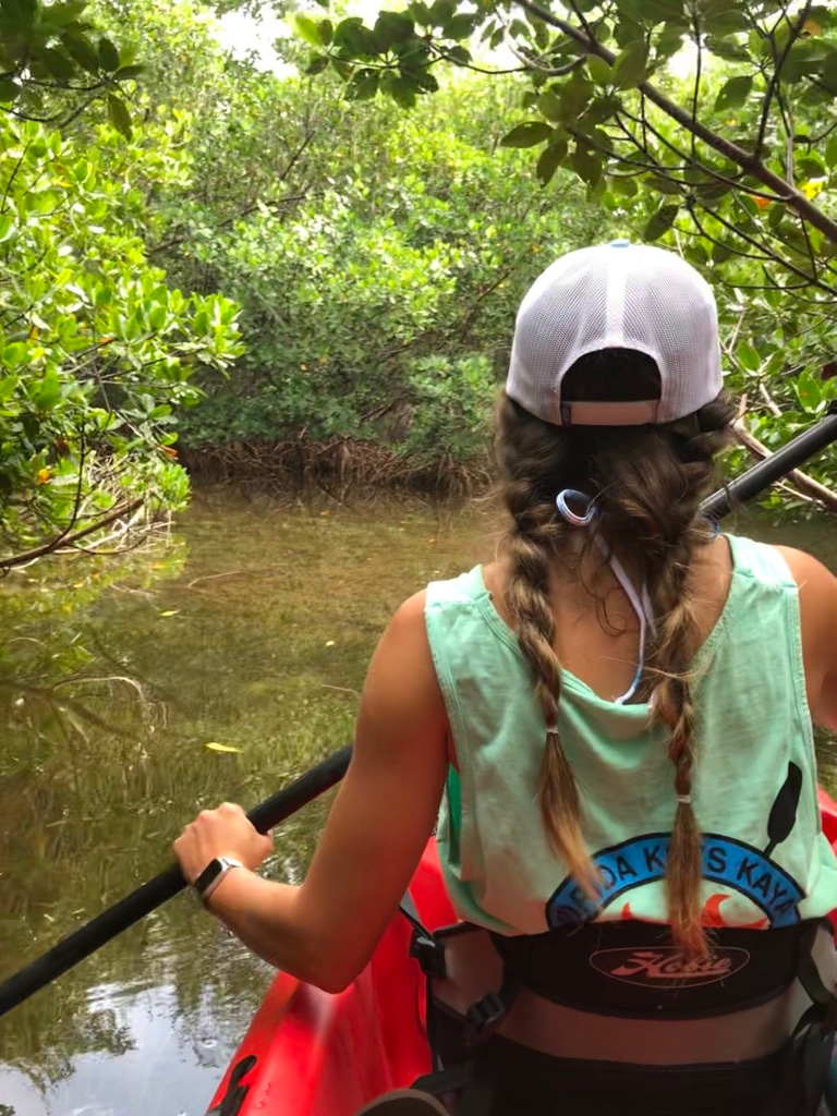 Person kayaking through lush mangrove forest, wearing a cap and tank top.
