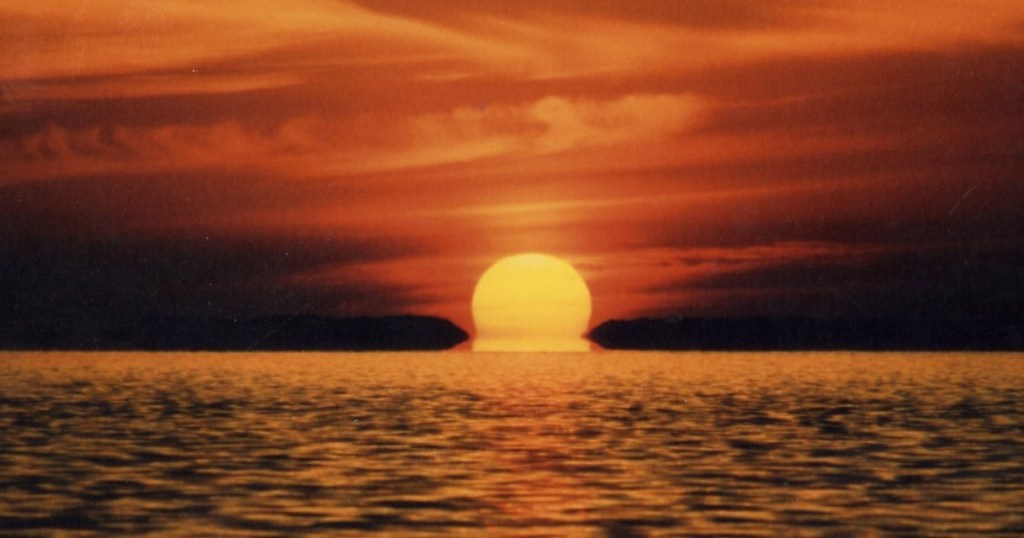 Sunset over the ocean with vibrant orange sky and reflection on water.