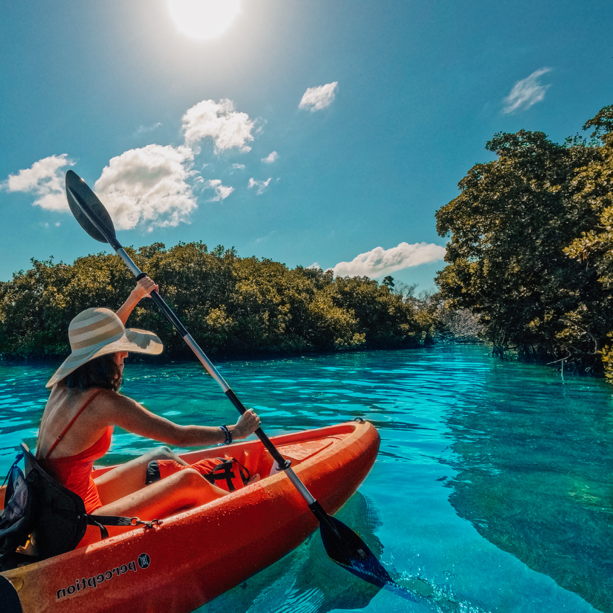Person in hat kayaking on clear blue water near dense green trees under sunny sky.