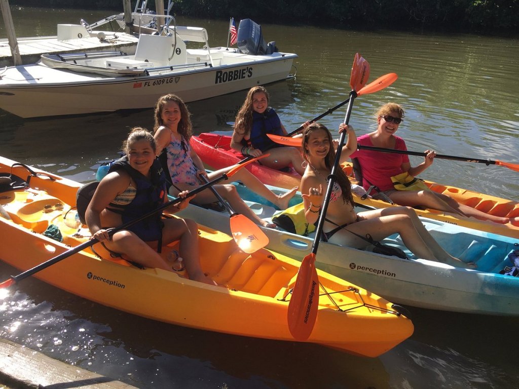 Group of smiling people in kayaks on a sunny day near a docked boat.