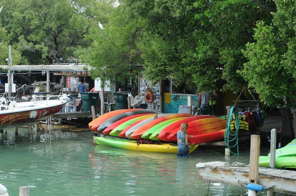 Colorful kayaks stacked by waterfront with trees and boats nearby.